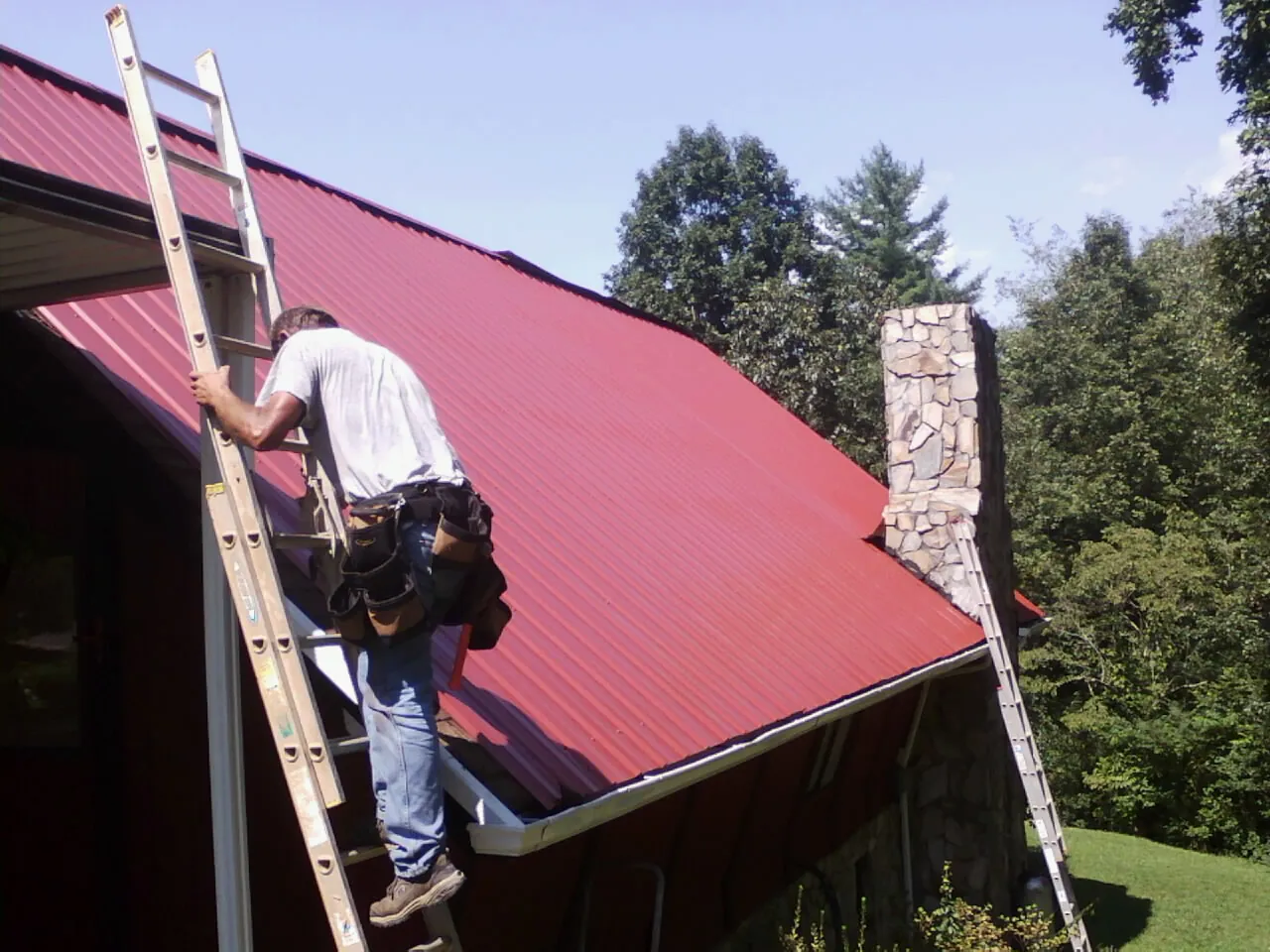 Roofer on a ladder beside a red metal roof and stone chimney — Soffit & Fascia Repair by Roofing Brothers in Parowan